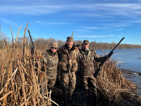 A group of men in camouflage standing in a wetland area, wearing camouflage clothing and holding guns.