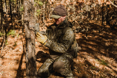 Wearing the Non-Typical Endurance Pant with Scent Control by Nontypical, a person in camouflage and yellow gloves kneels in a sunlit forest, attaching a trail camera to a tree among fallen leaves and tall pines.
