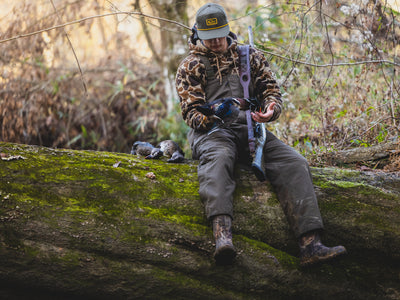 Wearing the Waterfowl LST Hardline Insulated Bib, a hunter sits on a mossy log in the woods, holding a bird and rifle, with two more birds beside them amid dense trees and foliage.