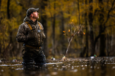 A bearded man wearing the LST BMF Jacket with Primaloft scans the sky in a flooded forest, highlighting its water-resistant, lightweight design for active hunting.