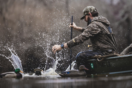 Man in camo waders leans over a boat with duck decoys nearby, wearing BMF EST Offset Quarter Zip Hoodie, ideal for waterfowl hunting with advanced features.