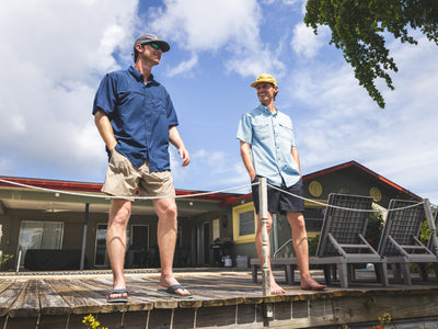 Two men wearing the 8-Shot Flyweight Shirt by Sale stand and smile on a sunny wooden deck outside a house, with lounge chairs and greenery in the background.