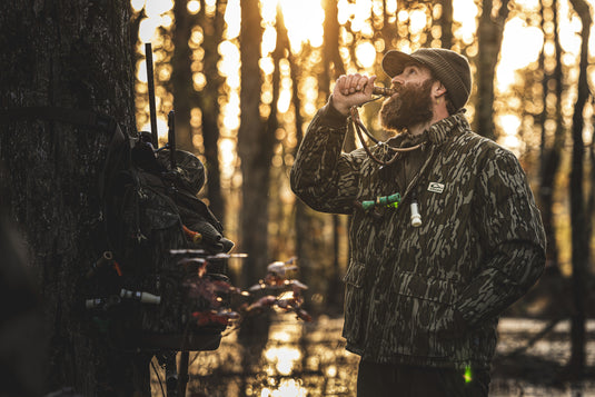 A bearded man in camouflage stands by a tree, blowing a duck call. His Waterfowl Throwback 80 Insulated Sport Bomber Jacket and hunting gear hang nearby as sunlight filters through the forest.