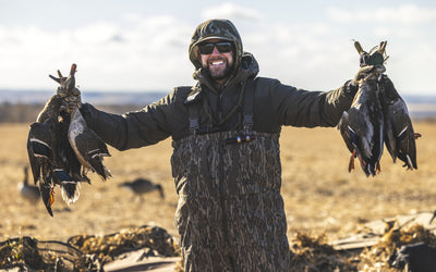 A smiling person in the Waterfowl LST BMF Pit Bib with Primaloft holds several ducks in each hand, standing outdoors in a field under a cloudy sky.