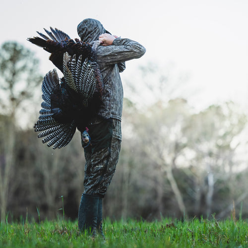 A man outdoors wearing a Turkey Bib with Spider Web Technology, featuring stretch material, cargo pockets, and water-resistant knee reinforcement, while carrying a turkey on his back.