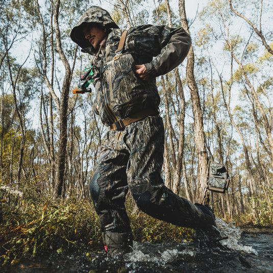 Wearing the Waterfowl BMF1600 Kevlar Wader, a person in camouflage strides through a forest stream, splashing water with each step.