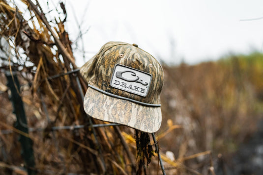 A Drake X Lost Hat Goat Rope Cap by Waterfowl hangs on a fence surrounded by dry, brown vegetation in an outdoor setting.