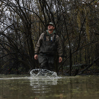 Wearing the Hardline Uninsulated Breathable Wader by Waterfowl, a person strides through knee-deep swamp water, splashing amid leafless trees in a forested area.