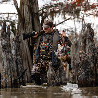 A hunter in Waterfowl Hardline Insulated Breathable Wader carries a rifle and ducks while trekking through a swamp wearing insulated, breathable waders and Thinsulate rubber boots.