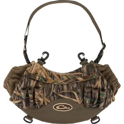 The Waterfowl LST Handwarmer Muff in Mossy Oak Original Shadow Grass features a windproof handwarmer design, insulated interior, shell holders, adjustable strap, and waterproof phone pocket. Displayed on a white background.