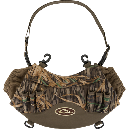 The Waterfowl LST Handwarmer Muff in Mossy Oak Original Shadow Grass features a windproof handwarmer design, insulated interior, shell holders, adjustable strap, and waterproof phone pocket. Displayed on a white background.