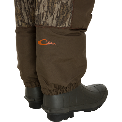 A close-up of lower legs in brown camo hunting pants, tucked into boots, with an orange logo on the right leg—perfect gear for a secure Youth Neoprene Wader fit by Waterfowl. Adjustable belt included.