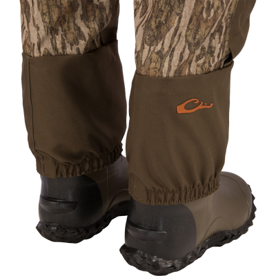 Hardline Breathable Youth Wader with camo fabric, reinforced knees, elastic cuffs, and black rubber boots, featuring an orange logo and cargo pouch for outdoor protection.