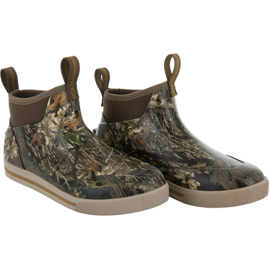 The Waterfowl Camp Deck Boot features an ankle-high, waterproof design with Mossy Oak Original Break Up camo, brown pull tabs, and tan rubber soles—perfect for outdoor use.