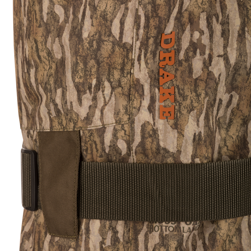 Close-up of Hardline Insulated Breathable Wader, showcasing camo fabric, adjustable strap with buckle, and orange DRAKE logo, highlighting durable, waterproof, insulated features.