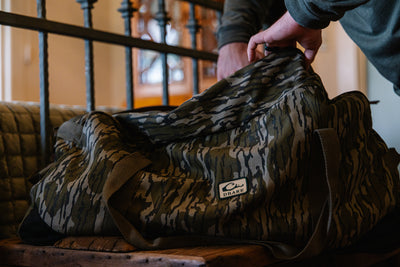 A person opens a Waterfowl Throwback 80 Duffle Bag with a camouflage pattern and Drake logo, resting on a wooden bench near a quilted cushion and iron railing.