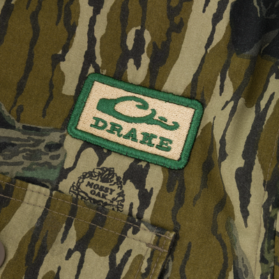 The Campfire Canvas Bomber Jacket by Lifestyle features rugged green and brown camo, a rectangular Drake patch, and a small logo above the Mossy Oak-labeled pocket.
