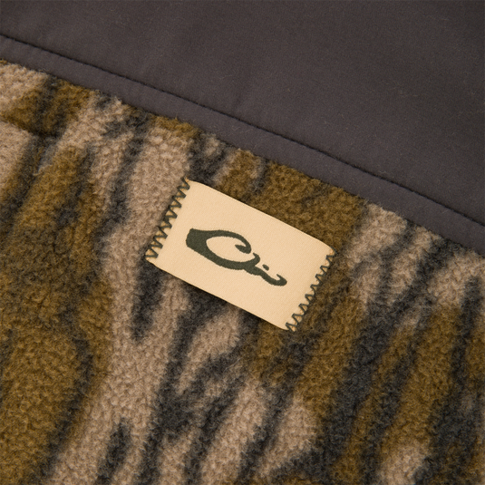 Close-up of a tan fabric tag featuring a black duck silhouette logo, stitched onto the olive and brown camouflage water-resistant fleece of a Camp Fleece Over Under Quarter Zip.