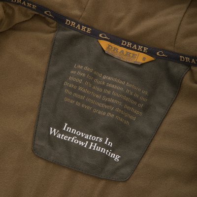 Close-up of the LST BMF Jacket with Primaloft, highlighting the inner pocket with Drake label and printed text about waterfowl hunting heritage and innovative design.