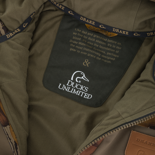 Interior view of a Drake X Ducks Unlimited LST BMF Jacket with Primaloft showing the neck label printed with a Ducks Unlimited logo and descriptive text about waterfowl conservation heritage.