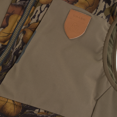 Close-up of a Drake X Ducks Unlimited LST BMF Vest with Primaloft, showing a tan chest panel with a leather-style shield patch embossed with a duck logo and the words Drake Ducks Unlimited.