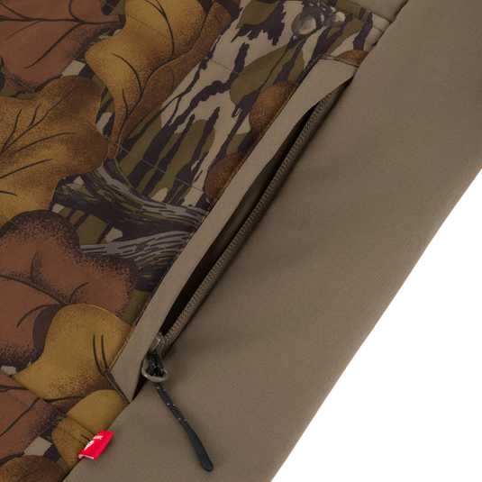 Close-up of a brown camouflage Drake X Ducks Unlimited LST BMF Vest with Primaloft featuring a zippered pocket and a small red tag on the lower seam.