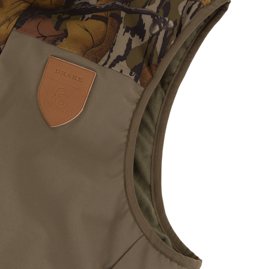 Olive green Drake X Ducks Unlimited LST BMF Vest with Primaloft featuring a camouflage shoulder panel and a leather patch embossed with a duck logo and the words Drake and Ducks Unlimited.