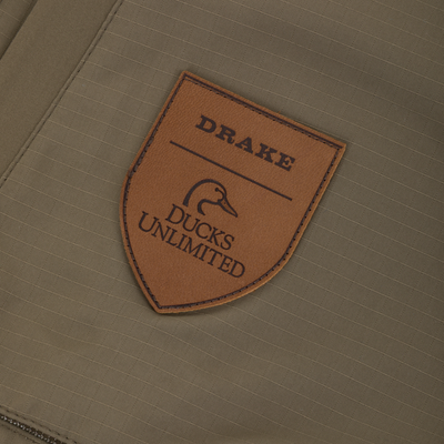 Brown shield-shaped leather patch featuring the Drake logo and Ducks Unlimited text, stitched onto the olive green fabric of a Drake X Ducks Unlimited LST BMF Pit Bib with Primaloft.