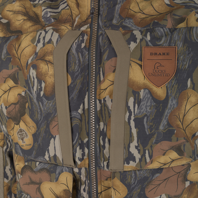 Close-up of a Drake X Ducks Unlimited LST BMF 3-in-1 Pit Jacket with Primaloft camouflage jacket with a front zipper, tan trim, and a leather patch featuring a duck logo.