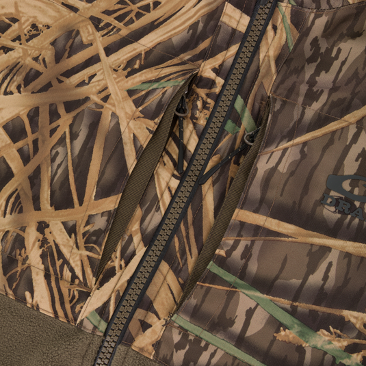 Close-up of the CommandXT Eqwader Vest by Waterfowl, an abrasion-resistant camo vest with a brown zipper and pocket featuring tan, green, and brown grass and branch patterns. Brand logo partially visible on right side.