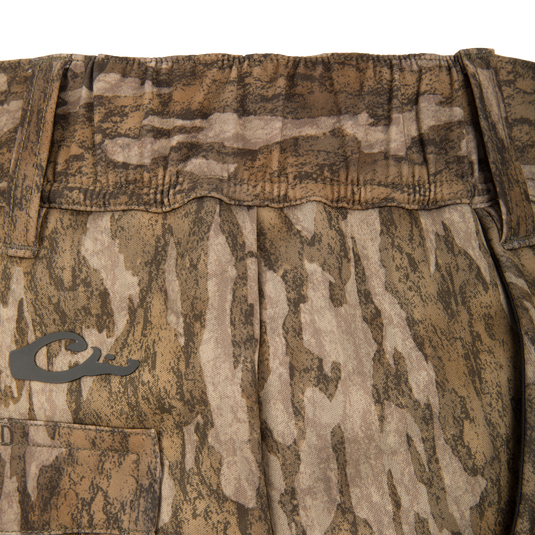 Close-up of Waterfowl BMF Ultimate Wader Pants shows camouflage-patterned fabric, a small pocket, stylized C logo, and soft polyester fleece shell at the waistband and upper section.