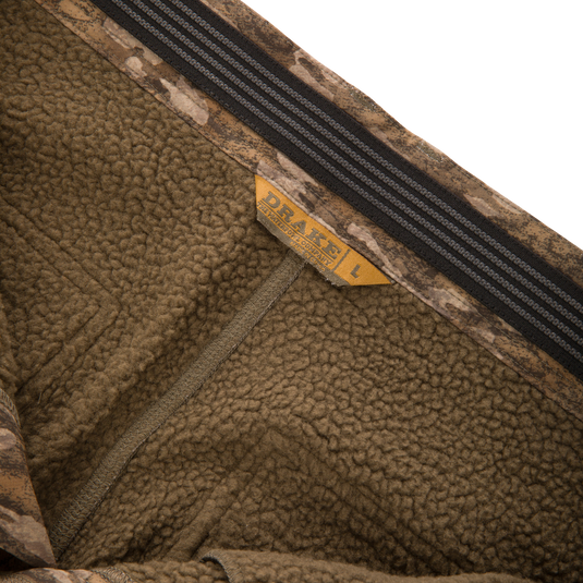Close-up of the inside of BMF Ultimate Wader Pants from Waterfowl, showing brown fleece with camouflage trim, yellow L size tag, soft polyester shell, Sherpa lining, and black-gray stripe detail.