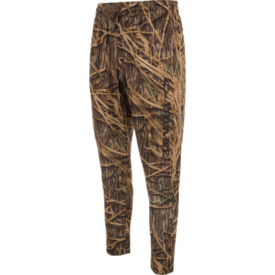 Waterfowl MST Under-Wader Jogger Pant features a brown and green tall grass camo pattern, elastic waistband, drawstring, tapered legs, and HUK logo—ideal waterfowl sweatpants. Shown on a white background.