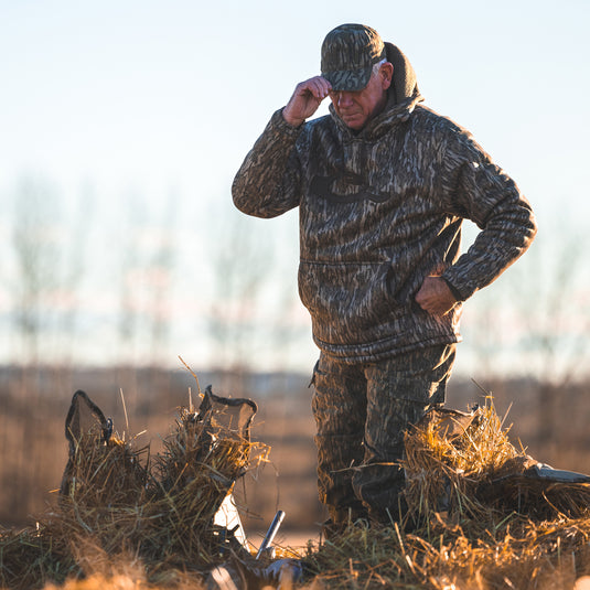 A man in camouflage LST Silencer Fleece-Lined Hoodie, featuring a double-lined hood and kangaroo pouch, ideal for outdoor activities. Made of polyester microfiber interlock fabric with fleece backing.