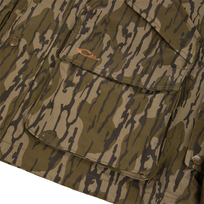 Close-up of a green and brown camouflage MST Drake Dry Waxed Cotton Wading Jacket, featuring a large front cargo pocket with an embroidered logo, metallic snaps, and a durable waterproof construction.