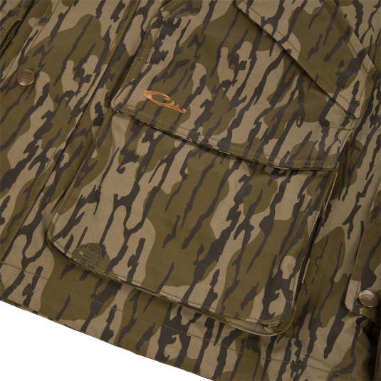 Close-up of a green and brown camouflage MST Drake Dry Waxed Cotton Wading Jacket, featuring a large front cargo pocket with an embroidered logo, metallic snaps, and a durable waterproof construction.