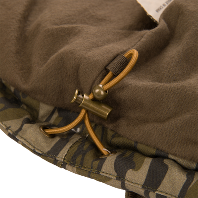 Close-up of the Waterfowl LST Insulated Waxed Cotton Jacket hem featuring an adjustable elastic drawstring cord with a metal toggle fastener against a brown fleece lining and camouflage waxed cotton exterior.