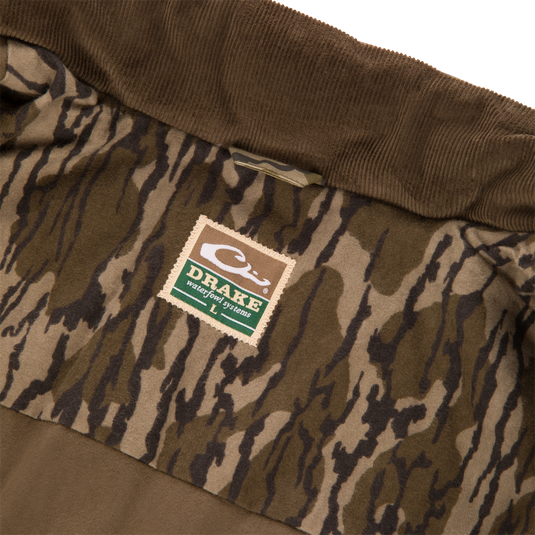 Interior of a Waterfowl LST Insulated Waxed Cotton Jacket featuring a brown corduroy collar and a camo-print lining with a brand label reading Drake Waterfowl Systems and size L.