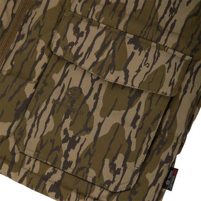 Close-up of a LST Insulated Waxed Cotton Vest pocket in brown and green camo by Waterfowl, featuring a flap, detailed stitching, small logo, and black tag at the lower edge.