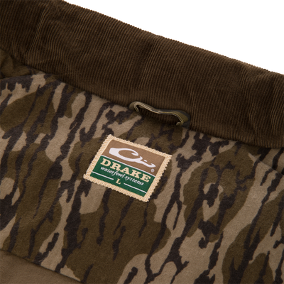The LST Insulated Waxed Cotton Vest by Waterfowl features a brown corduroy waistband, camouflage fabric, a green and tan Drake Waterfowl Systems label, and a metal hook above the label.
