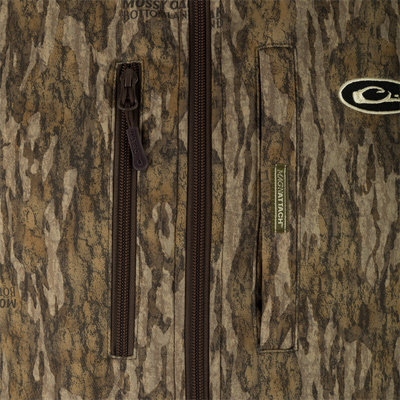 Close-up of a camouflage-patterned jacket with brown zippers, a visible Mossy Oak Bottomland logo, and a MAGNATACH green label on the right side, layered over a Waterfowl MST Hole Shot Two-Tone Eqwader Vest.