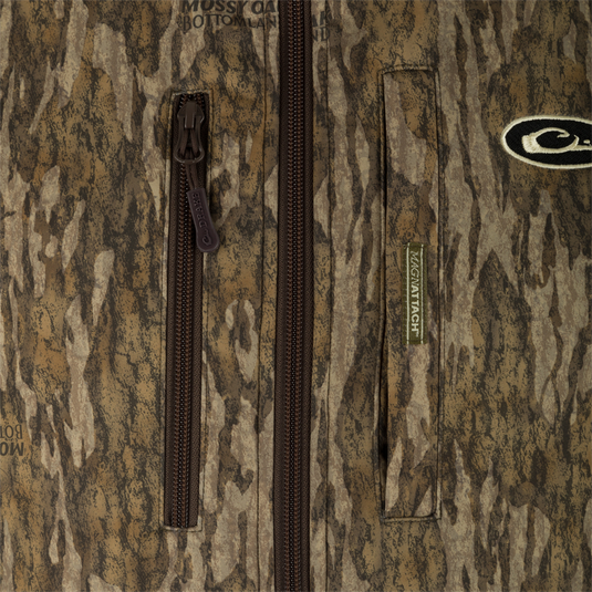 Close-up of a camouflage-patterned jacket with brown zippers, a visible Mossy Oak Bottomland logo, and a MAGNATACH green label on the right side, layered over a Waterfowl MST Hole Shot Two-Tone Eqwader Vest.
