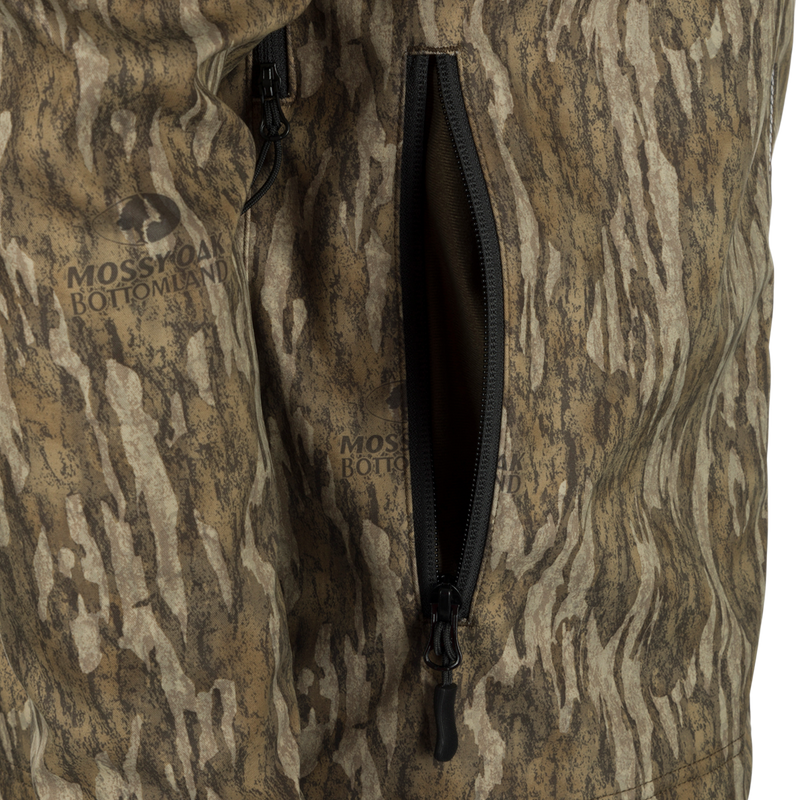Close-up of MST Windproof Softshell Jacket with Mossy Oak Bottomland camo, showcasing a partially unzipped pocket and logo, highlighting its hunting functionality.