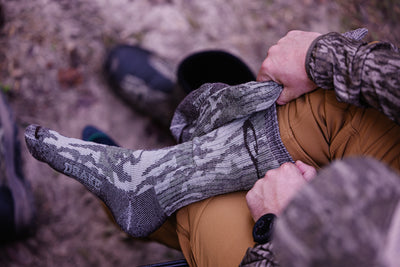 A person outdoors pulls up a Midweight Merino Camo Mid Calf Hunting Sock over their foot, showcasing its camo pattern and durable design.