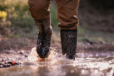 Person walking through puddle in camouflage EST Uninsulated 16 Hybrid Mudder Boot 3.0, showcasing water-resistant durability and comfort for rugged environments.