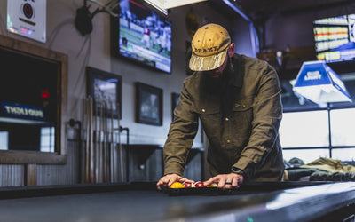 Man in Delta Moleskin Shirt Jacket, featuring reinforced forearms and brass snaps, sets up billiard balls in a bar scene, reflecting rugged outdoor style.