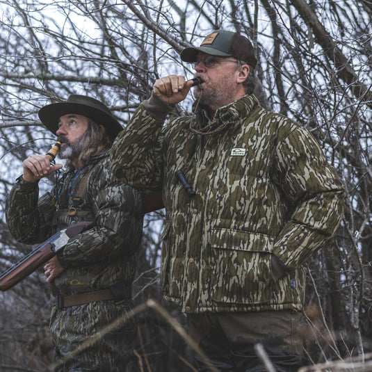 Two people in camo, one wearing a Waterfowl Throwback 80 Featherless Puffer Jacket, use duck calls in the woods. One holds a shotgun and wears a wide-brimmed hat among bare branches on a cloudy day.