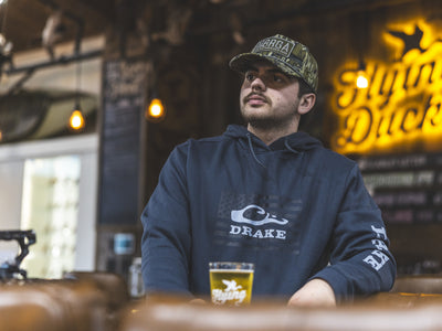 Young man in a Drake Cotton Logo Work Hoodie, sitting in a taproom, showcasing its comfort and durability amidst warm Edison lights.