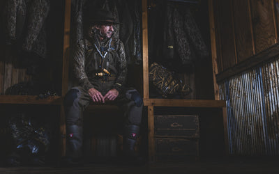 A man in camouflage hunting gear and a Waterfowl EST BMF Offset 1.0 Crew sits pensively on a bench in a dimly lit wooden room, surrounded by coats, boots, and equipment.