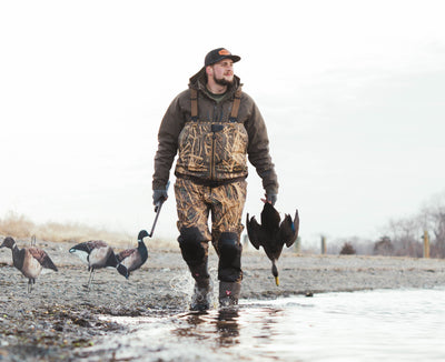 Wearing the Waterfowl LST CommandXT 3-in-1 Primaloft Insulated Jacket, a man in camo waders stands by the water with decoys and a duck, under a cloudy sky in a sparse landscape.